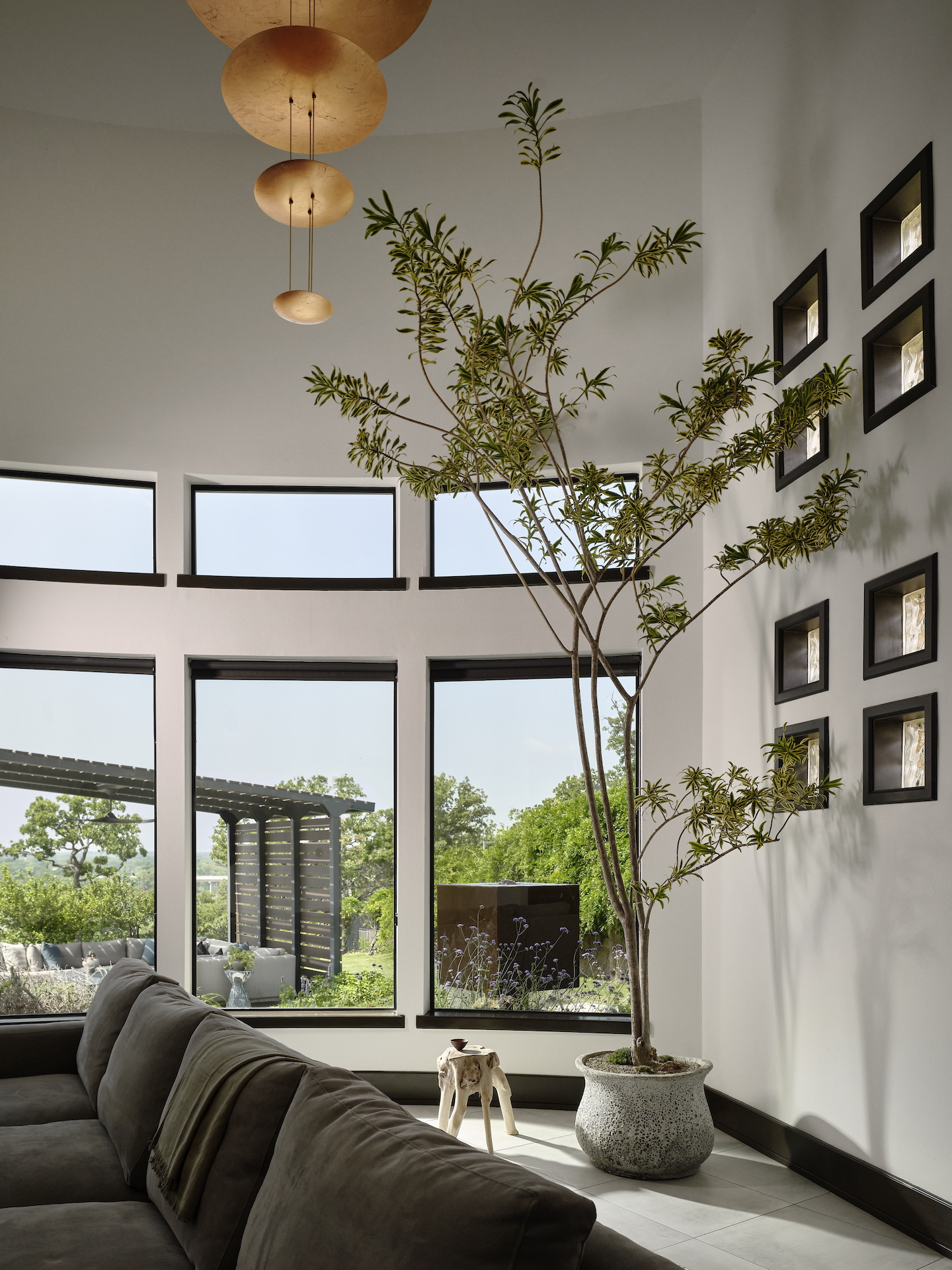 A modern living room with a large potted tree, gray sectional sofa, pendant lights, and tall windows offering views of greenery outside. Small framed art is arranged on the white wall.