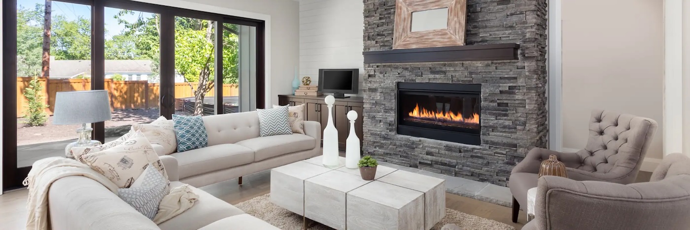 Modern living room with light sofas, a stone fireplace, large windows, and a view of a fenced backyard. Neutral decor and natural light fill the space.
